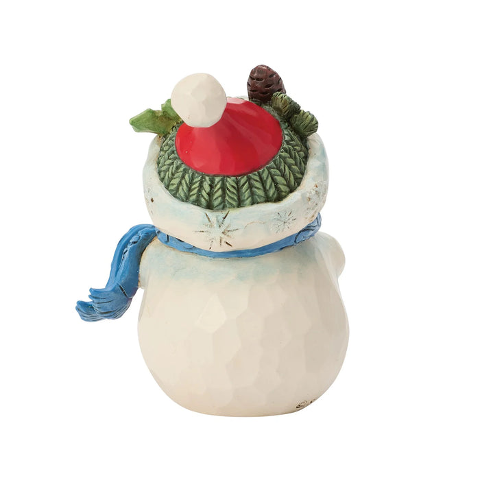 A small ceramic snowman figurine with a textured off-white body, a blue scarf, a green knit hat, a red top, and a small brown pinecone on top, isolated on a white background.