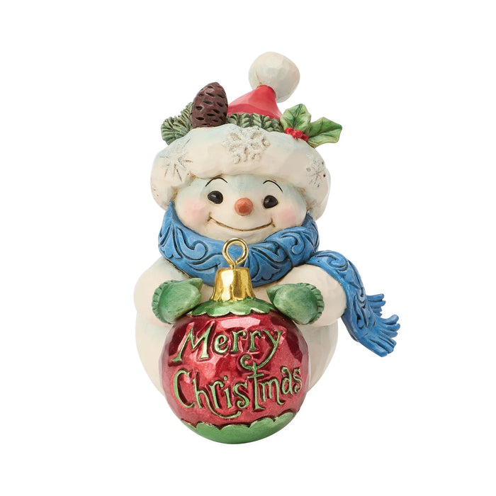 Snowman figurine with a hat and blue scarf holding a green and red ornament that says "Merry Christmas" on a white background