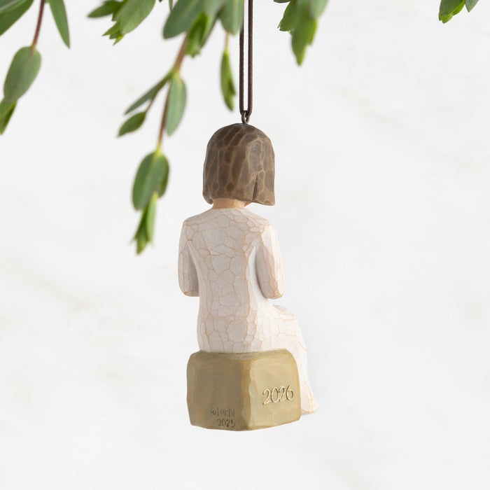 Decorative figurine of a woman sitting on a block with '2026' on it, hanging from a branch.