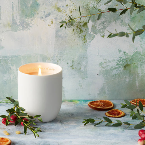 White candle in a textured container with dried oranges and greenery on a light background