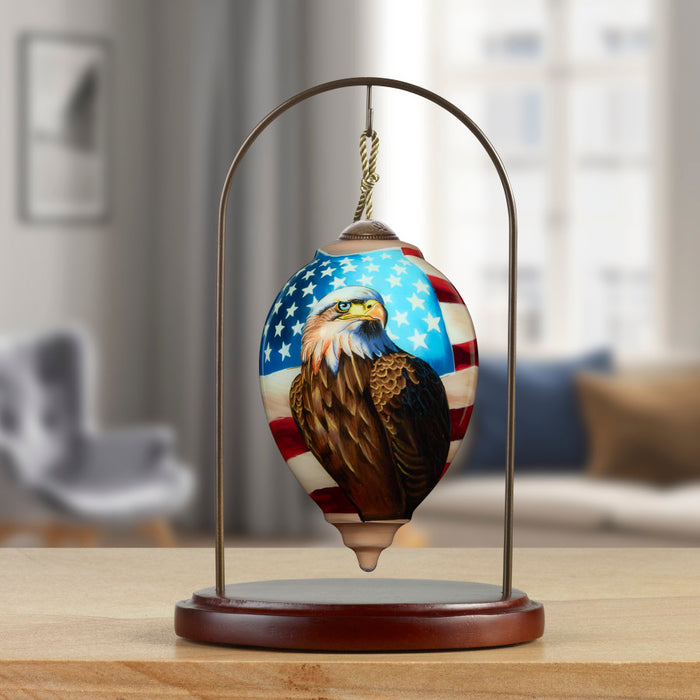 A decorative hanging ornament shaped like a teardrop, featuring a painted bald eagle over an American flag design, suspended from a metal stand with a wooden base on a tabletop. The background shows a softly blurred living room interior.