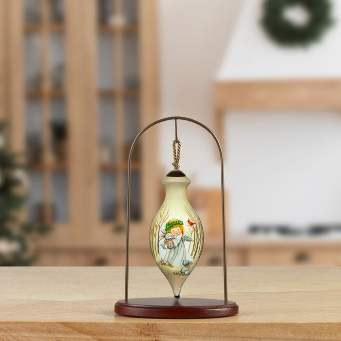 A small decorative ceramic ornament or vial with a painted winter scene, displayed on a metal hanger stand with a wooden base on a tabletop. The background is a softly blurred indoor room with holiday decor.