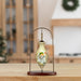 A small decorative ceramic ornament or vial with a painted winter scene, displayed on a metal hanger stand with a wooden base on a tabletop. The background is a softly blurred indoor room with holiday decor.
