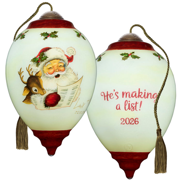 A Christmas ornament shown from both sides on a plain white background. One side features a painted Santa Claus reading with a reindeer and holly accents, and the other side reads 'He's making a list! 2026' in red script.