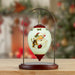 A Christmas ornament displayed on a metal stand, featuring a hand-painted Santa Claus and reindeer illustration on a glossy white teardrop-shaped ornament with red accents, set against a softly blurred festive background.