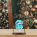 A Christmas ornament shaped like a teardrop, featuring a family of snowmen painted on a light blue background, hanging from a brass stand with a dark wooden base on a wooden tabletop.