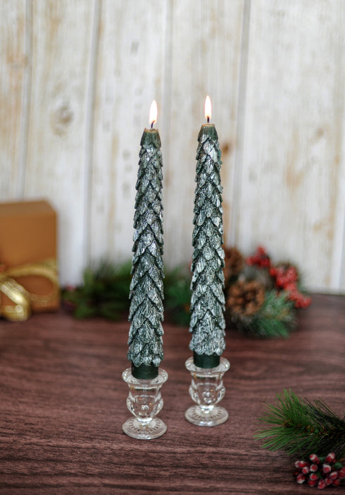 Two green textured candles in glass candlesticks on a wooden surface with festive decorations.