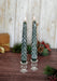 Two green textured candles in glass candlesticks on a wooden surface with festive decorations.