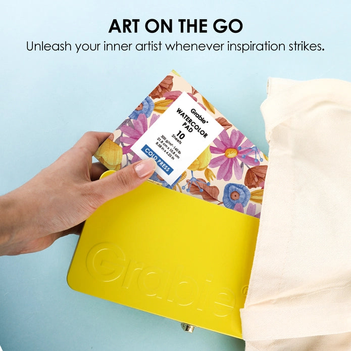 Hand holding a colorful watercolor pad with a yellow holder against a light blue background.