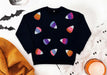 A black crewneck sweatshirt decorated with colorful glittery candy corn motifs, presented in a flat-lay arrangement with a textured orange blanket, a white lace runner, and Halloween-themed props.