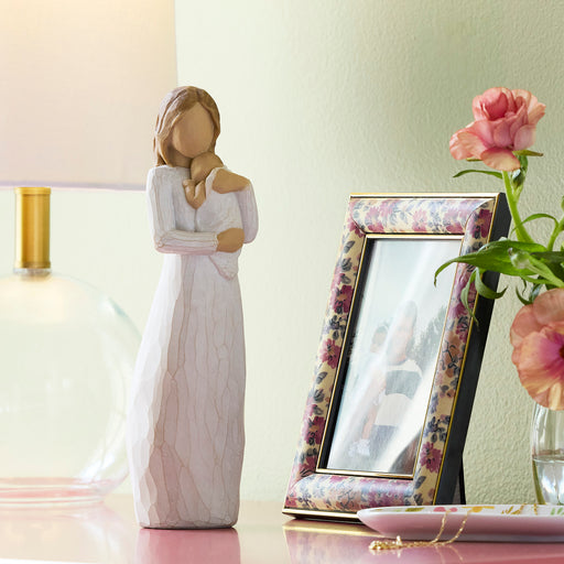 Decorative figurine of a woman in a white dress next to a floral photo frame and pink flowers on a table.
