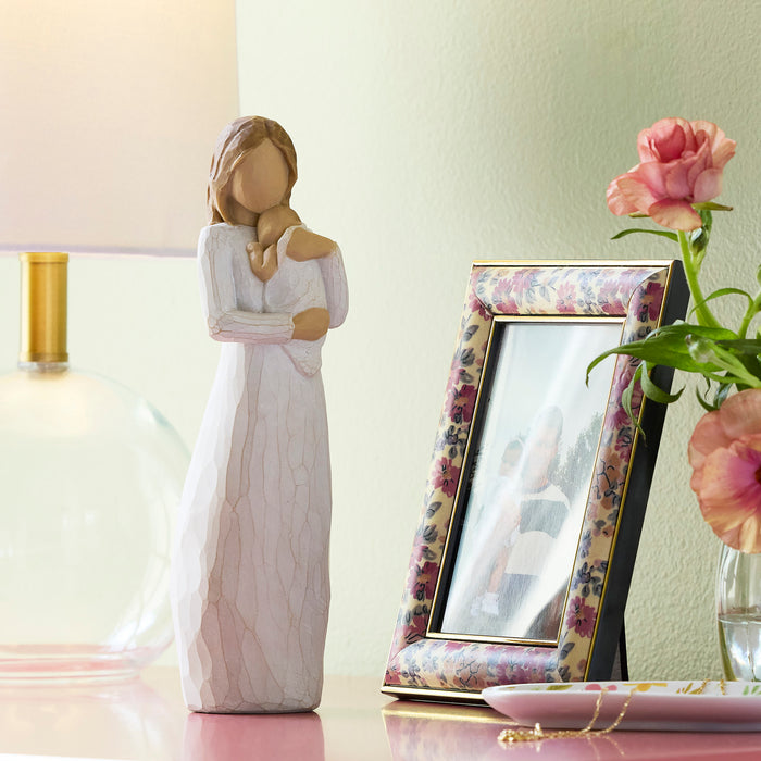 Decorative figurine of a woman in a white dress next to a floral photo frame and pink flowers on a table.
