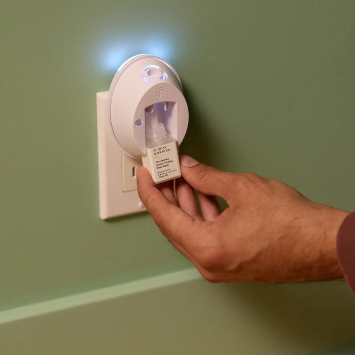 A hand inserts a battery into a white circular plug-in device mounted on a light green wall, with a small blue light glowing from the device.