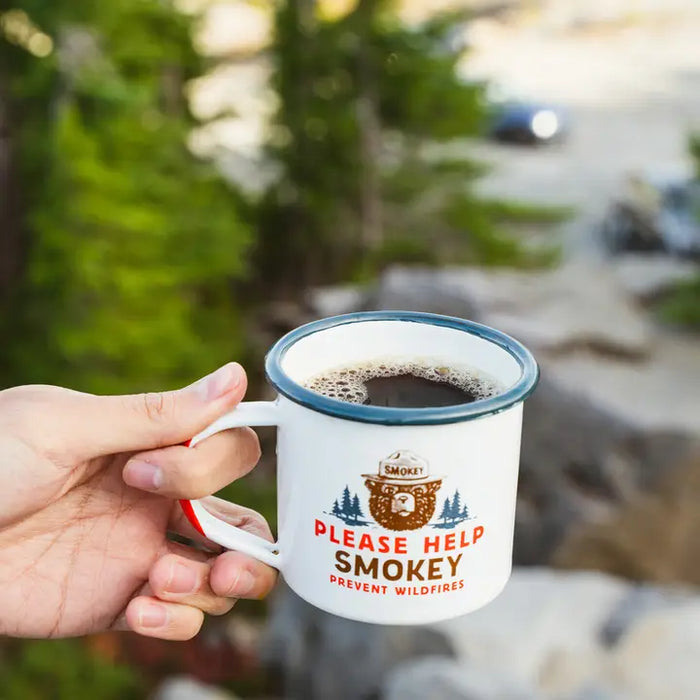 Person holding a Smokey Bear coffee mug outdoors with trees in the background