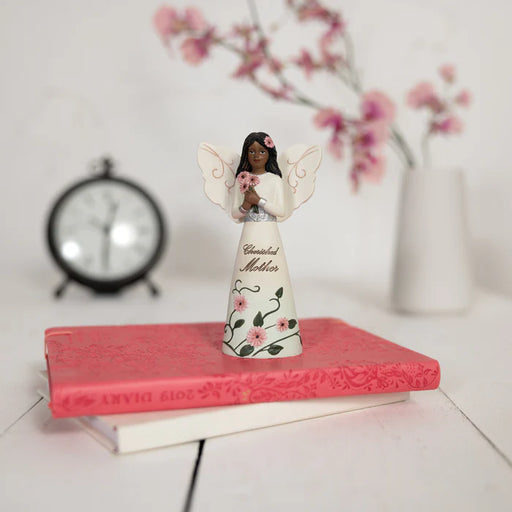 Decorative angel figurine on a pink book with a clock and flowers in the background