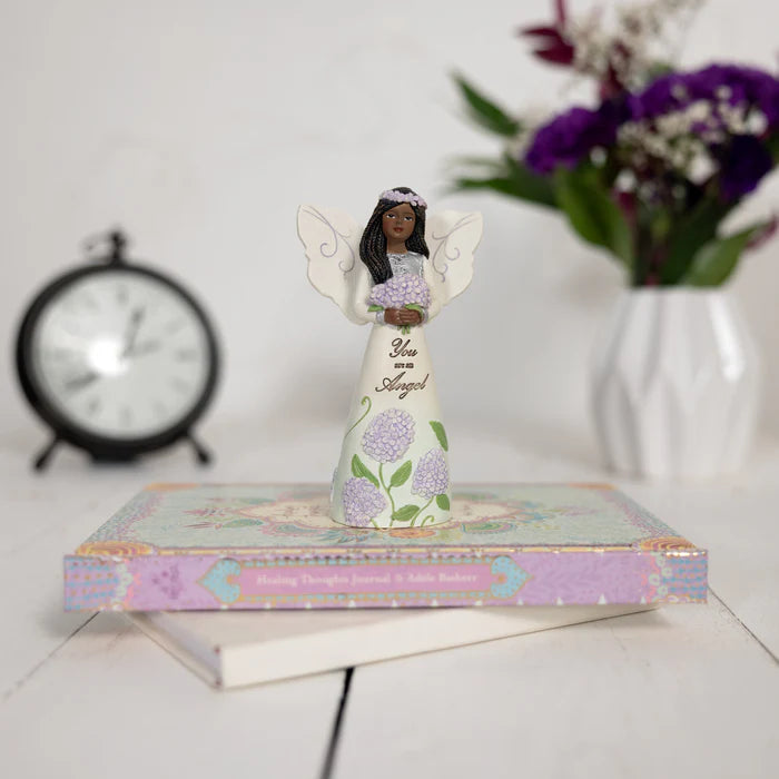 Decorative angel figurine with flowers on a book, with a clock and flowers in the background.