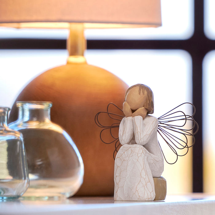 Wooden angel figurine with wire wings on a surface with a blurred lamp and glass jars in the background.