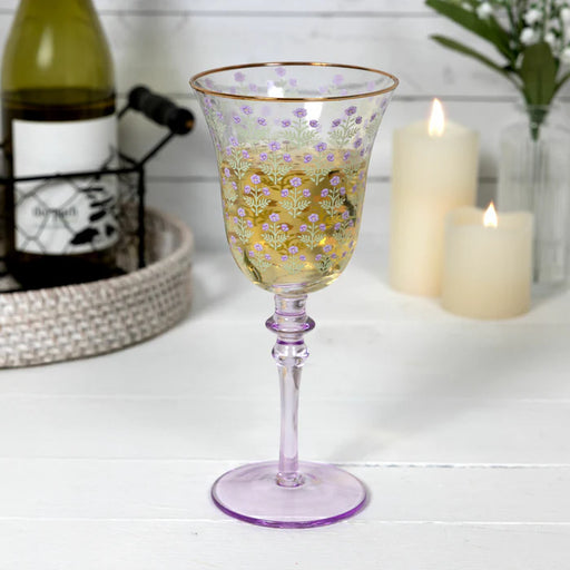 Decorative wine glass with floral pattern on a white surface with candles and a bottle in the background.