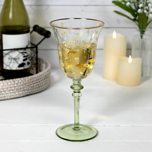 Green glass goblet with floral design filled with a clear liquid, bottle, and candles in the background on a white surface.