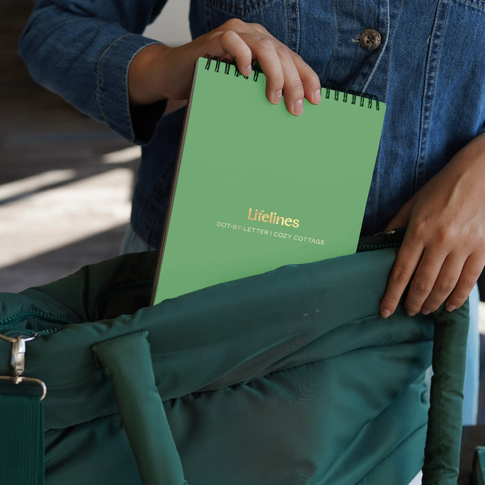 Person holding a green notebook titled 'Lifelines' with a denim jacket and green bag in the background.