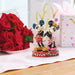 Figurines of a cartoon couple on a decorative stand with red roses and a heart-shaped gift in the background.