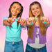Two smiling young women holding up small colorful ring-shaped accessories toward the camera against a pink background.