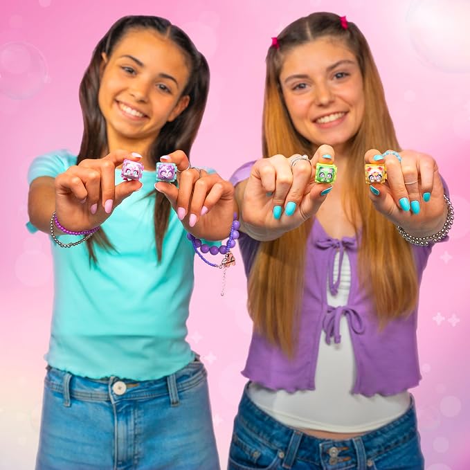 Two smiling young women holding up small colorful ring-shaped accessories toward the camera against a pink background.
