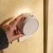 Hand holding a small cylindrical light fixture against a wall with floral patterns.