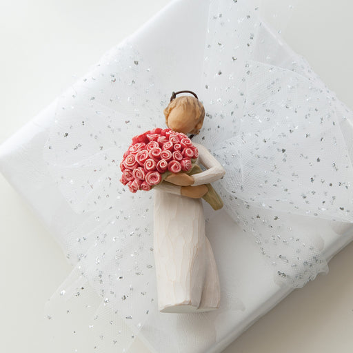 Small figurine of a person holding a bouquet of pink flowers on a textured white surface.