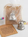Silver bell keychain with engraved text, placed on a white surface with a sheer bag and card.