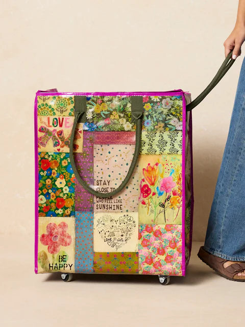 Multicolored floral-patterned rolling bag with text held by a person wearing blue jeans.