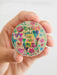 Hand holding a colorful round sticker with floral designs and text on a white background