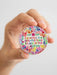 Hand holding a colorful circular sticker with text and floral design on a white background