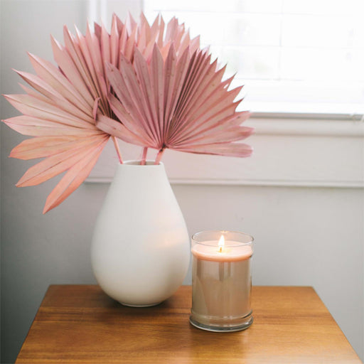 White vase with pink dried leaves and a lit candle on a wooden surface.