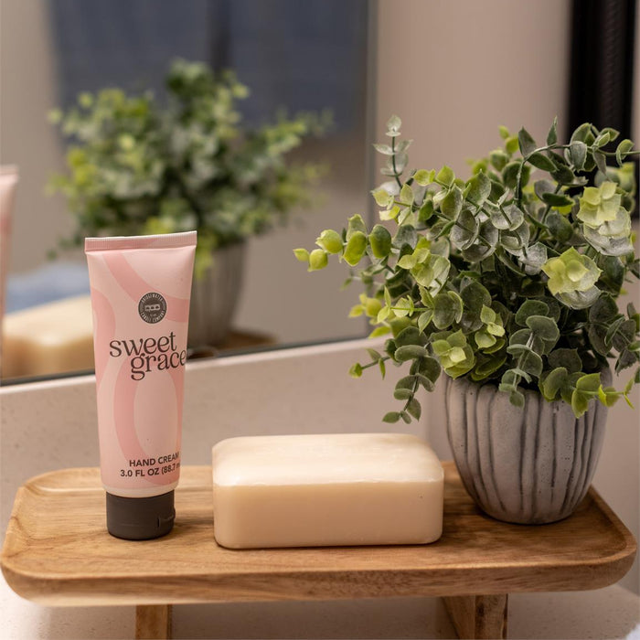 Bathroom setting with a pink tube of 'sweet grace' hand cream, a bar of soap, and a potted plant on a wooden tray.