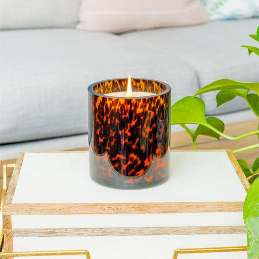 Tortoiseshell-patterned candle in a glass holder on a marble surface with a light gray sofa and plant in the background.