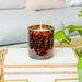 Tortoiseshell-patterned candle in a glass holder on a marble surface with a light gray sofa and plant in the background.