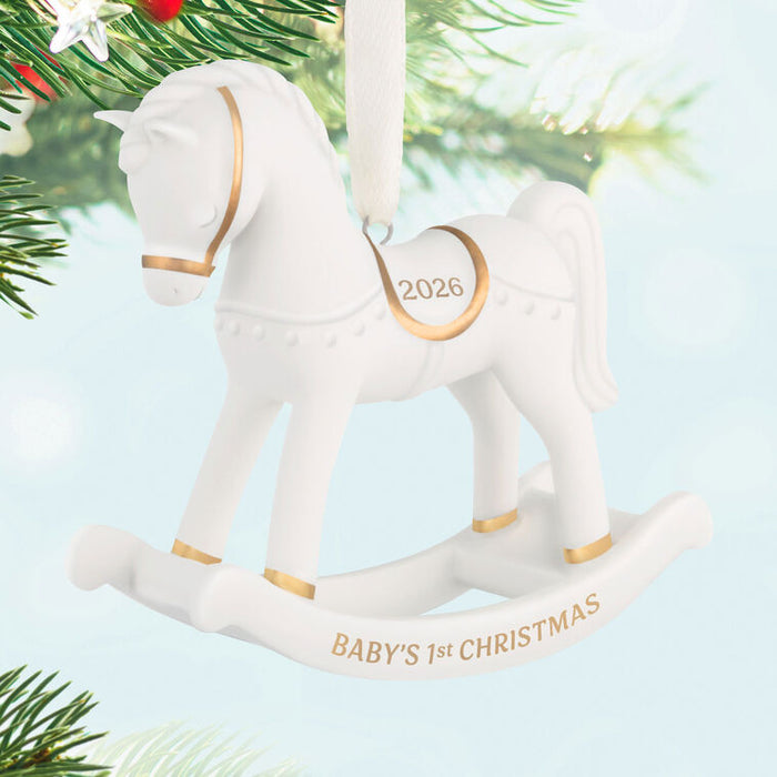 White rocking horse ornament with gold accents, '2026' and 'BABY'S 1ST CHRISTMAS' text, on a Christmas tree.