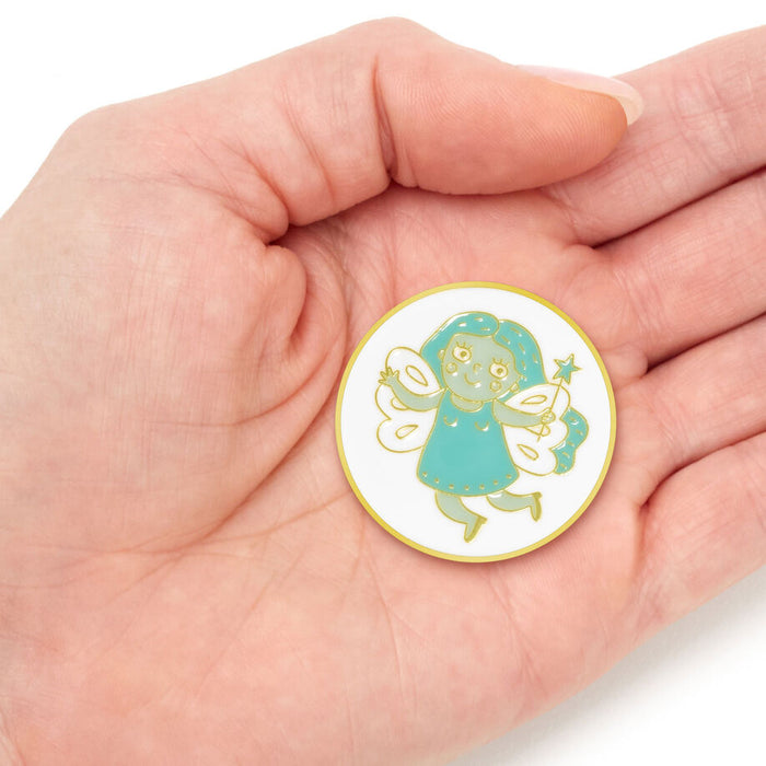 A small round enamel pin featuring a cute green fairy character with white wings and a teal dress, displayed on an open hand against a white background.