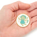 A small round enamel pin featuring a cute green fairy character with white wings and a teal dress, displayed on an open hand against a white background.