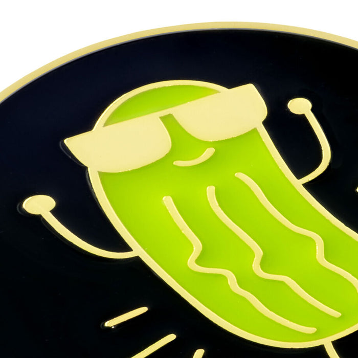 A close-up of a circular black enamel pin featuring a bright green pickle character wearing pale yellow sunglasses, with simple yellow line details and a gold-toned border.