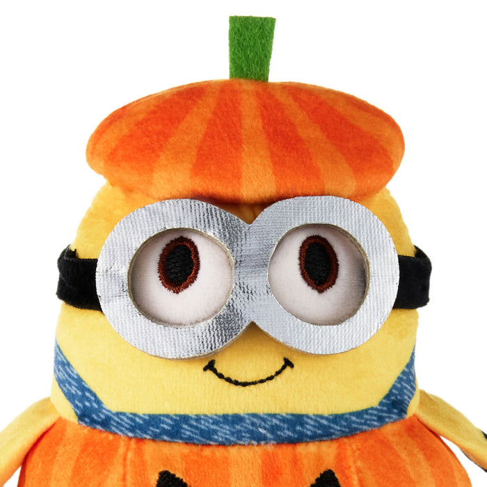 A plush Minion character dressed as a pumpkin, featuring an orange striped beret, large silver goggles, a blue denim-style scarf, and a yellow body on a white background.