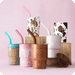 A group of patterned and printed tumblers with matching straws arranged on wooden blocks against a soft pink background. The tumblers include turquoise, pink, brown, white, and cow-print designs, with one featuring a cowboy and cactus motif.