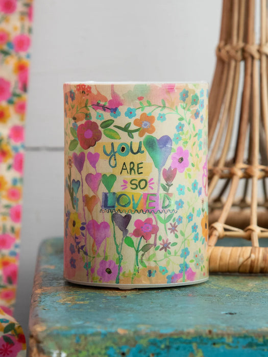 Decorative candle with floral design and 'You are so loved' text on a rustic surface.