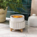 White ceramic planter with decorative stand on a marble surface, with a plant and other decor items in the background.