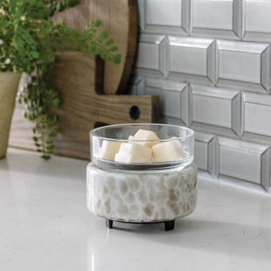 Ceramic wax warmer with glass container on a white surface, with a plant and wooden cutting board in the background.
