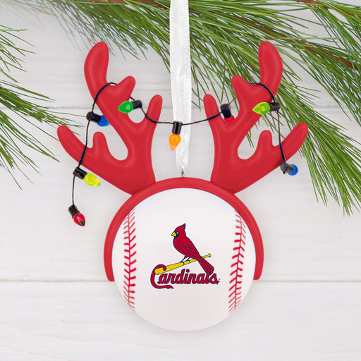 A Christmas ornament shaped like a baseball with red reindeer antlers and hanging multicolor lights, featuring the St. Louis Cardinals logo on the front, displayed against a white wood background with pine branches.