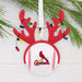 A Christmas ornament shaped like a baseball with red reindeer antlers and hanging multicolor lights, featuring the St. Louis Cardinals logo on the front, displayed against a white wood background with pine branches.