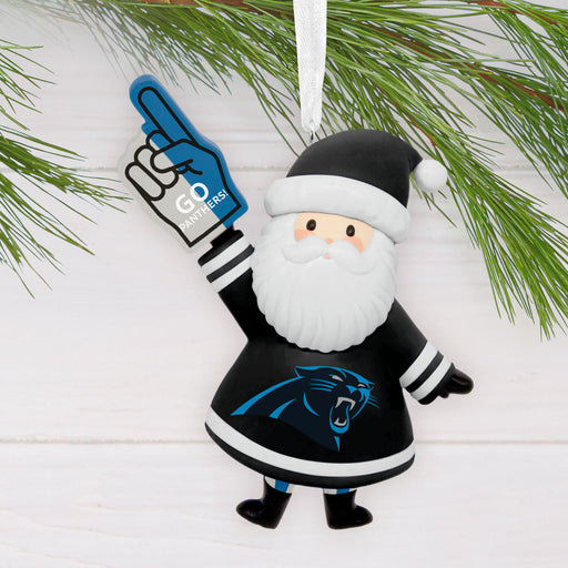 A Carolina Panthers Santa Claus hanging ornament styled with pine branches on a white wood background. The ornament depicts Santa in a black and white uniform with the team logo, holding a blue foam finger that says 'GD Panthers'.