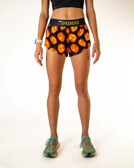 A model from the waist down wearing black athletic shorts with a vibrant orange and yellow pumpkin print, featuring the brand name 'CHICKANUFFS' on the waistband, paired with green sneakers against a white studio background.